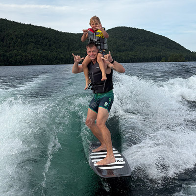 Man on wakeboard behind boat holding child on shoulders